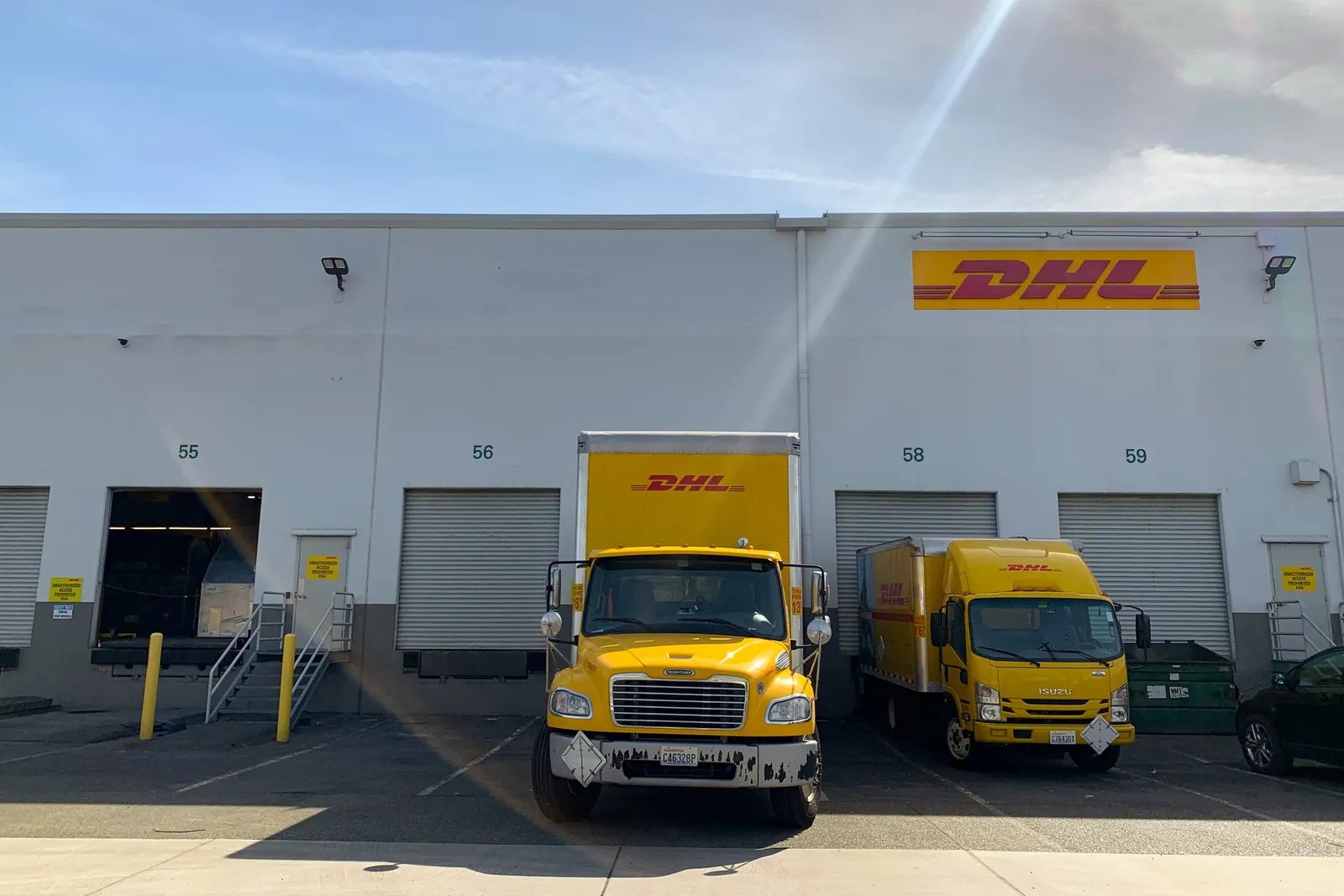 Two DHL-labeled trucking vehicles sit outside of warehouse doors.