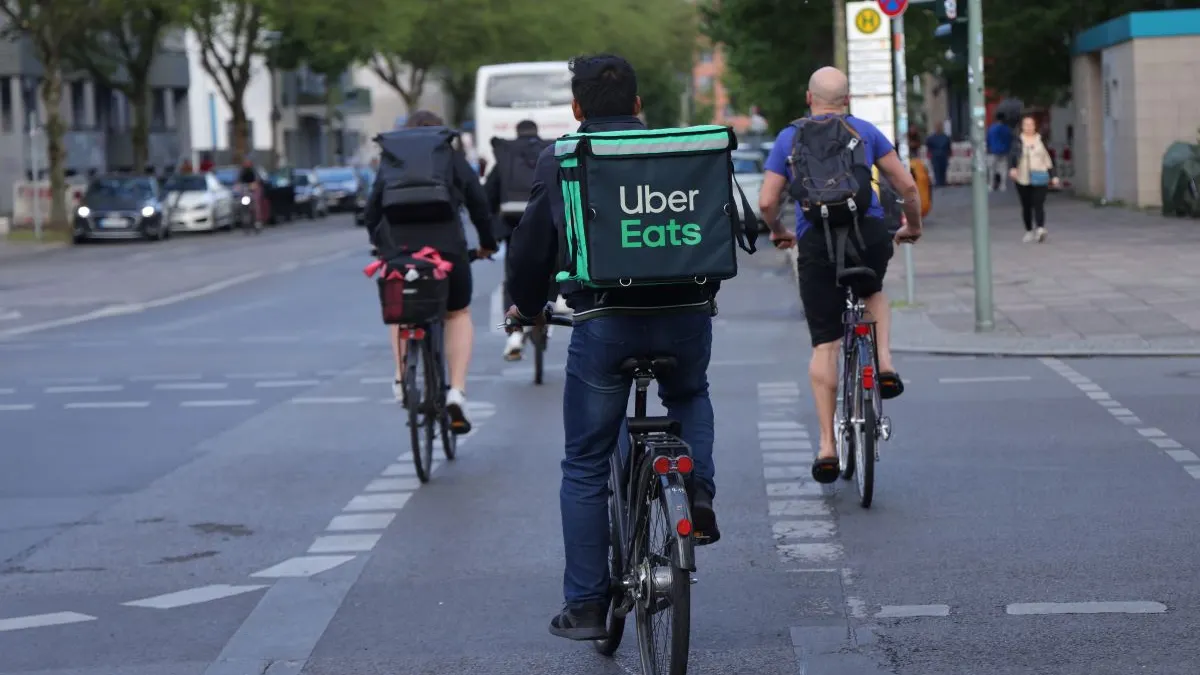 An image of four bike riders won a street. The bike rider in the foreground is carying an Uber Eats bag on his back.
