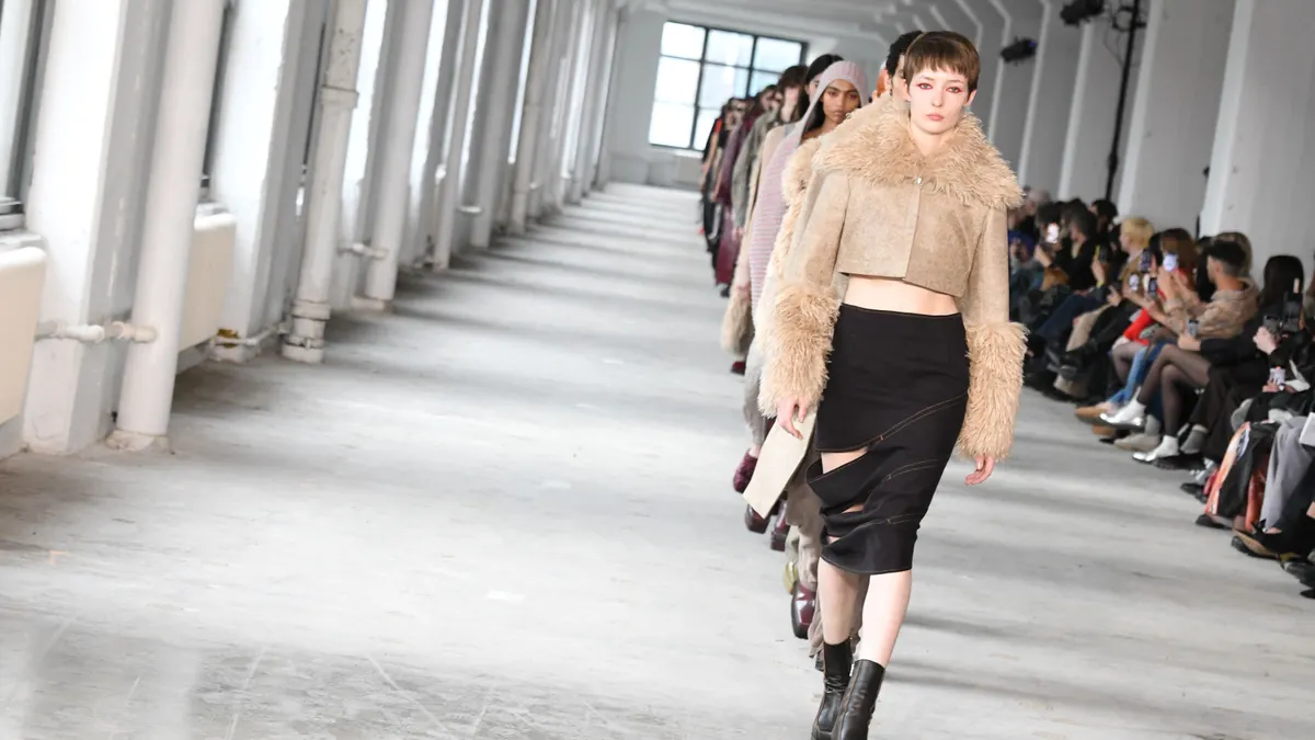 Models in jackets and coats walk down a runway as a seated crowd watches.